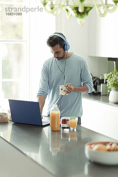 Mann mit Kopfhörern arbeitet von zu Hause aus am Laptop in der Morgenküche