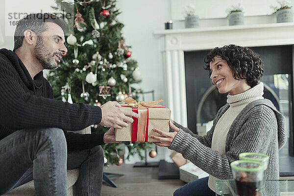 Ehemann gibt Weihnachtsgeschenk an Frau im Wohnzimmer mit Baum
