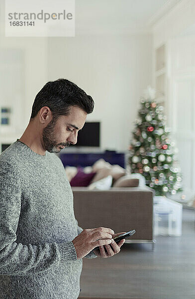 Mann mit Smartphone im Wohnzimmer mit Weihnachtsbaum