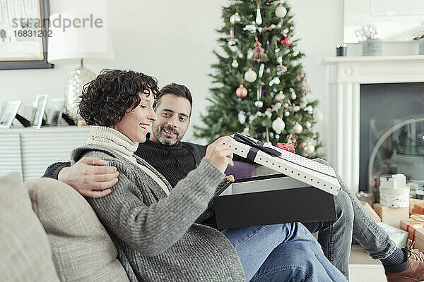 Frau öffnet Weihnachtsgeschenk vom Ehemann auf dem Wohnzimmersofa