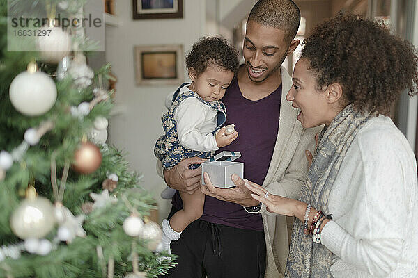 Glückliche Eltern helfen Baby Tochter öffnen Weihnachtsgeschenk durch Baum