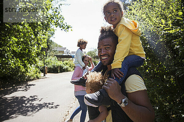 Porträt glücklicher Vater trägt Töchter auf sonnigen Bürgersteig