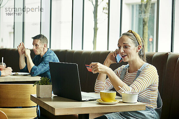 Frau sitzt in einem Café  benutzt einen Laptop und spricht mit einem Smartphone