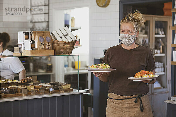 Kellnerin mit Gesichtsmaske bei der Arbeit in einem Café  trägt Teller mit Essen.