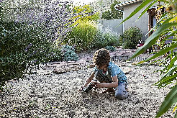 7 Jahre alter Junge spielt im sandigen Garten mit seinem Spielzeugschiff.