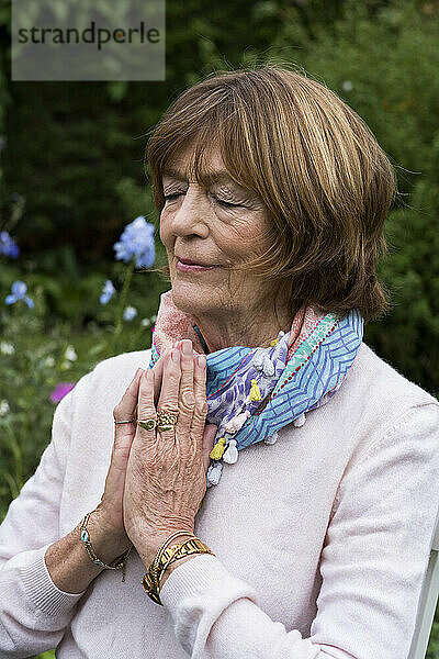 Frau mit zusammengelegten Händen und geschlossenen Augen sitzend in einem Garten