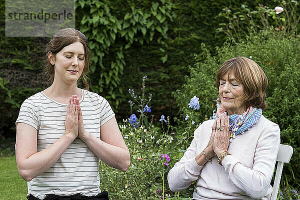 Frau und Therapeutin in einem Garten sitzend  Hände zusammen und Augen geschlossen.
