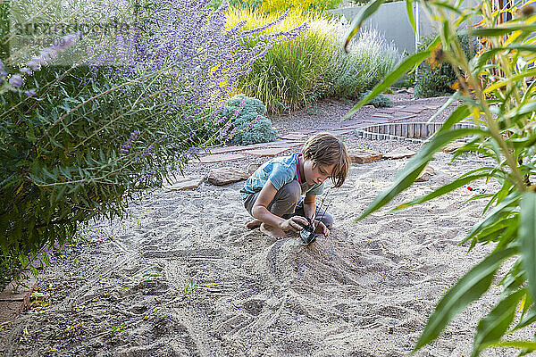 7 Jahre alter Junge spielt im sandigen Garten mit seinem Spielzeugschiff.