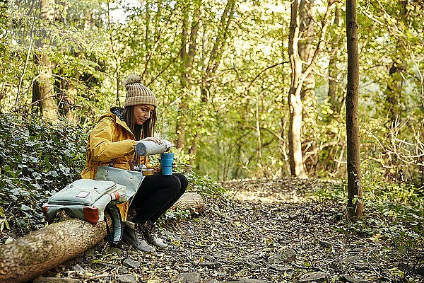 Frau sitzt auf Baumstamm im Wald gießt Getränk aus Thermoskanne