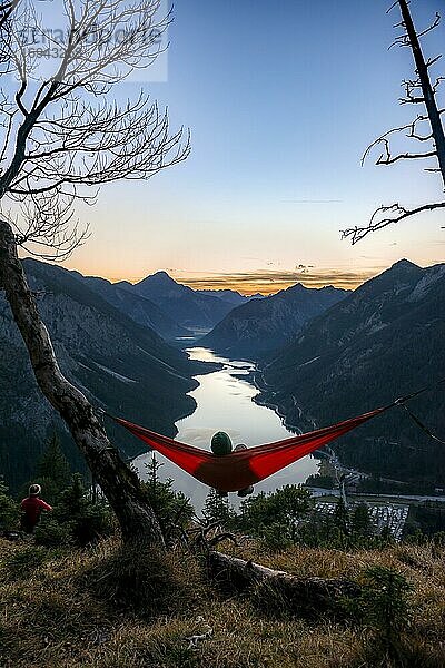 Junger Mann sitzt in einer roten Hängematte  Blick auf Berge mit See  Sonnenuntergang  Plansee  Tirol  Österreich  Europa