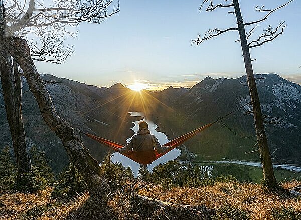 Junger Mann sitzt in einer roten Hängematte  Panoramablick auf Berge mit See  Sonnenstern  Sonnenuntergang  Plansee  Tirol  Österreich  Europa