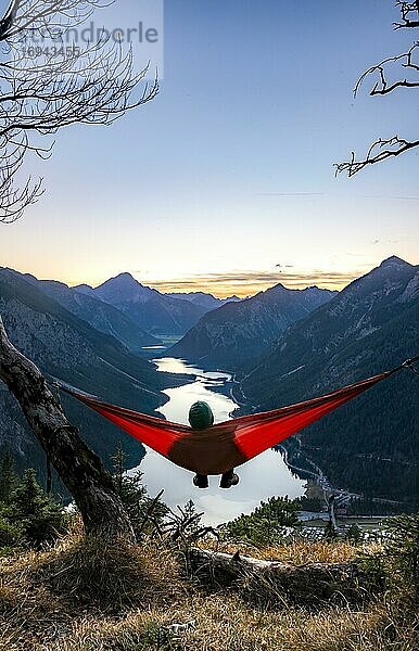 Junger Mann sitzt in einer roten Hängematte  Panoramablick auf Berge mit See  Sonnenuntergang  Plansee  Tirol  Österreich  Europa
