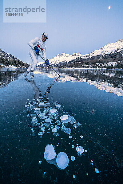 Eishockeyspieler Mann beim Schlittschuhlaufen auf dem mit Eisblasen bedeckten Silser See in der Abenddämmerung  Engadin  Kanton Graubünden  Schweiz  Europa