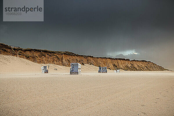 Strandkörbe am menschenleeren Strand von Kampen. Der stürmische Wind hat einen der Körbe umstürzen lassen. Im Hintergrund leuchtet das Rote Kliff in der tiefstehenden Sonne. Darüber wölbt sich ein bleigrauer Wolkenhimmel