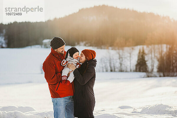 Nettes Baby und Eltern in verschneiten Bergen im Winter sonnigen Tag lächelnd