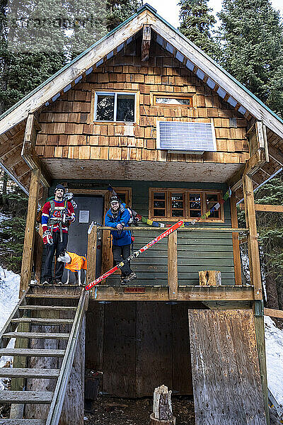 Junge Männer mit Eishockeyausrüstung  die lächelnd in die Kamera schauen  während sie auf der Veranda eines Holzhauses im Winter in British Columbia  Kanada  stehen