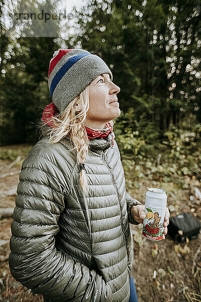 Lächelnde  glückliche blonde Frau in grauer Jacke und Wintermütze mit Bier in der Hand.