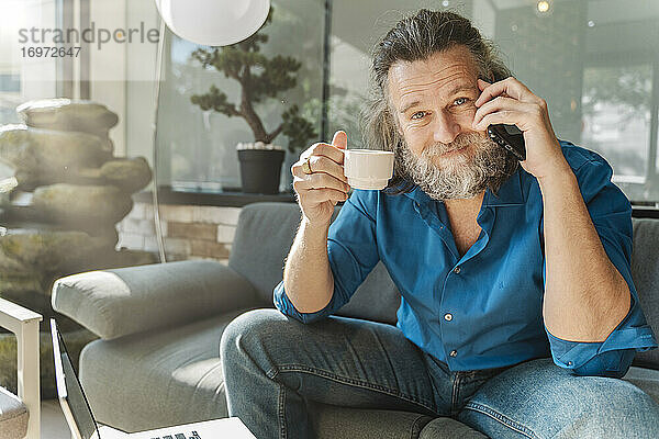 Älterer Mann trinkt einen Kaffee und telefoniert auf einem Sofa sitzend