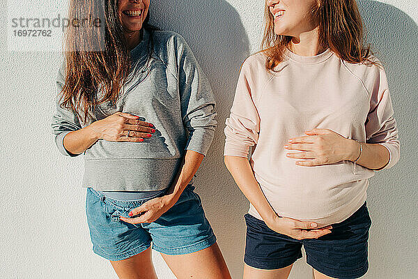 Zwei junge schwangere Frauen halten ihren Bauch und lächeln sich gegenseitig an
