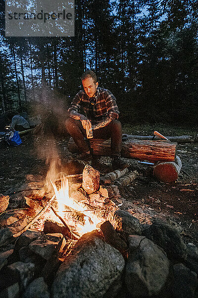 Junger Mann in Flanell mit Bier in der Hand sitzt nachts am Lagerfeuer in Maine