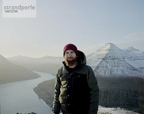 Mann schaut in die Kamera auf einem verschneiten Berggipfel mit Blick auf das Meer auf den Färöer Inseln