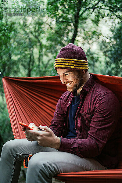 Junger Mann benutzt sein Smartphone in einer Hängematte im Wald