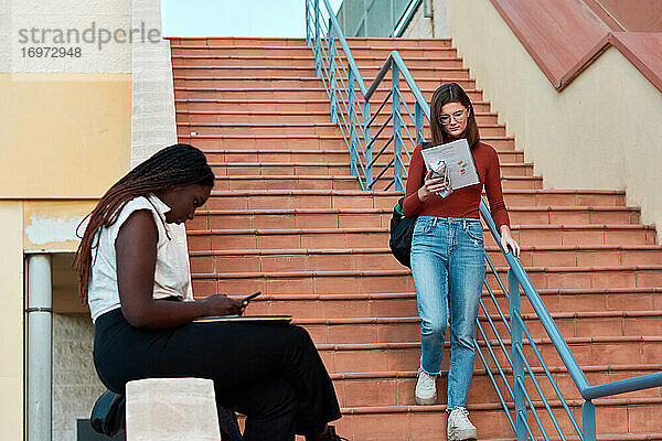 Eine Schülerin geht die Treppe hinunter  die andere schaut auf ihr Handy