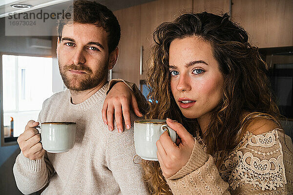Junges Paar mit heißem Getränk in der Küche zu Hause schaut in die Kamera