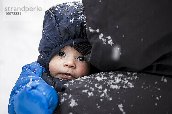 Bezauberndes Baby in warmer Kleidung in den Händen eines anonymen Elternteils bei einem Winterspaziergang bei verschneitem Wetter