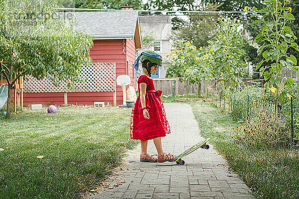 Ein Mädchen in rotem Kleid und Helm Schritte auf Skateboard im Garten im Sommer