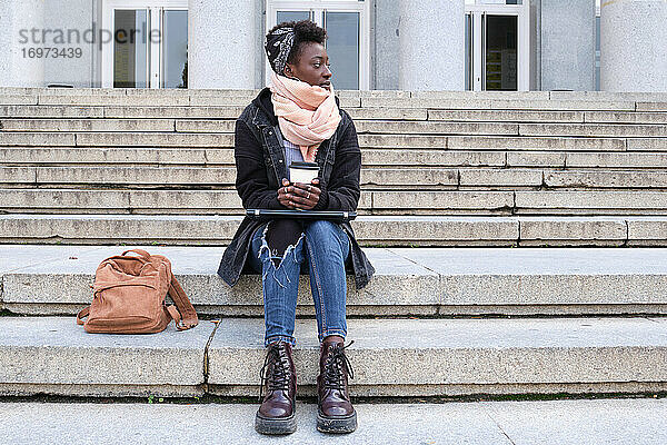Schöne Universität afrikanische Studentin Blick auf Seite  hält eine Tasse Kaffee  sitzen auf Treppen außerhalb auf dem Campus. College-Leben-Konzept.