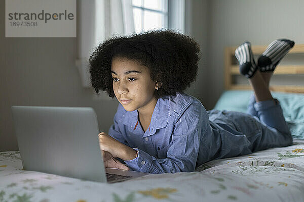 Zehnjähriges gemischtrassiges Mädchen arbeitet im Bett liegend an ihrem Laptop