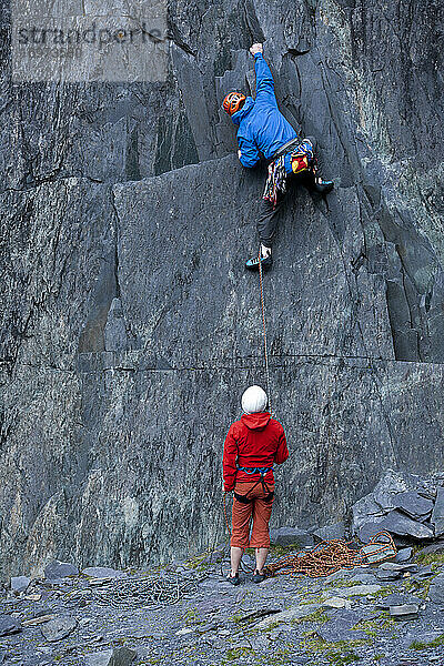Mann klettert in einem Schiefersteinbruch in Nordwales eine steile Felswand hinauf