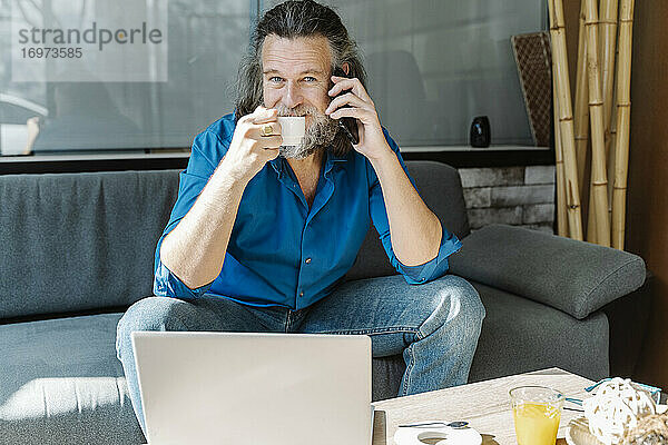 Älterer Mann mit Bart telefoniert und trinkt Kaffee sitzend