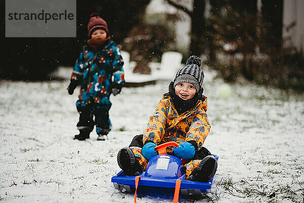 Junges Kind sitzt auf einem Schlitten und lächelt an einem verschneiten Tag im Hinterhof