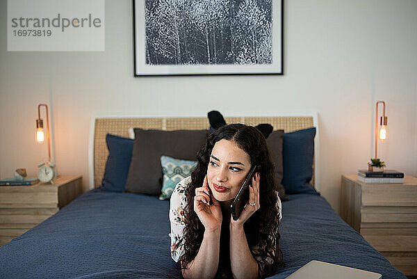 Glückliche junge Frau  die auf dem Bett liegt und mit einem Telefon spricht