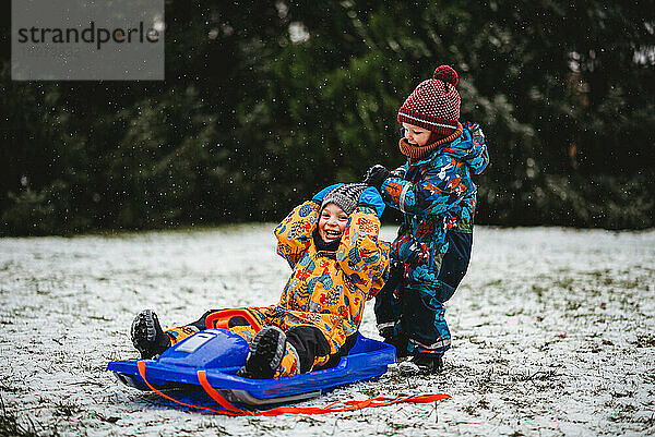 Junge Kinder spielen mit Schlitten im Schnee im Park mit Schneeanzügen und Mützen