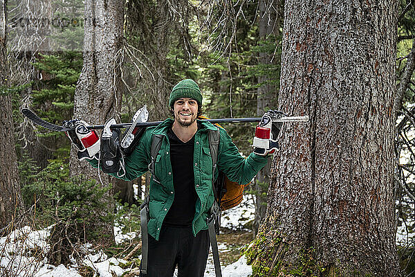 Fröhlicher junger Mann mit Hockeyschläger und Schlittschuhen auf den Schultern  der lächelnd in die Kamera schaut  während er in der Nähe von Bäumen im verschneiten Wald steht