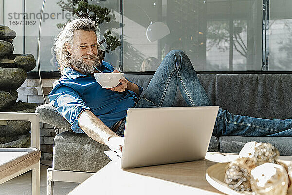 Älterer Mann mit Bart lächelt und arbeitet mit seinem Laptop auf einem Sofa