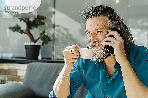Älterer Mann trinkt einen Kaffee und telefoniert auf dem Sofa in seinem Wohnzimmer. Business-Konzept