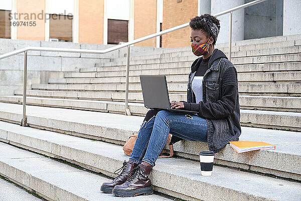 Afrikanische Studentin an der Universität  die einen Gesichtsschutz trägt und an ihrem Laptop arbeitet  während sie auf einer Treppe auf dem Campus sitzt. Neue Normalität im College.