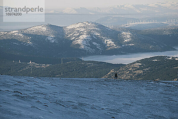 Mann mit Snowboard auf dem Rücken beim Wandern in der Sierra de Guadarrama vor Windmühlen und Berggipfeln  Madrid