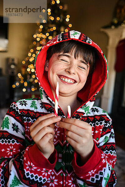 Fröhlicher Junge im festlichen Pyjama  der zur Weihnachtszeit eine Zuckerstange isst.