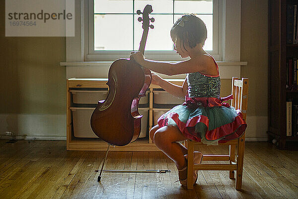 Ein kleines Mädchen in einem Tütü sitzt auf einem kleinen Stuhl am Fenster und hält ein Cello