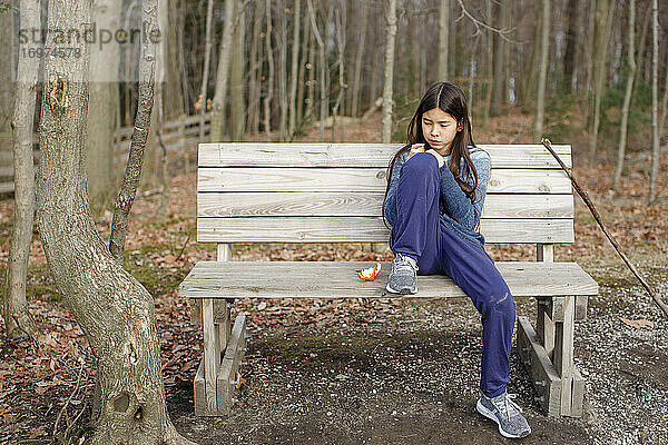 Ein kleines Mädchen sitzt allein auf einer Bank im Wald und isst eine Orange im Herbst