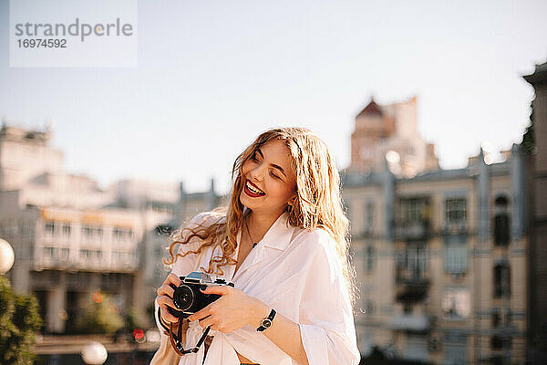 Glückliche junge Frau  die eine Kamera hält  während sie im Sommer in der Stadt steht