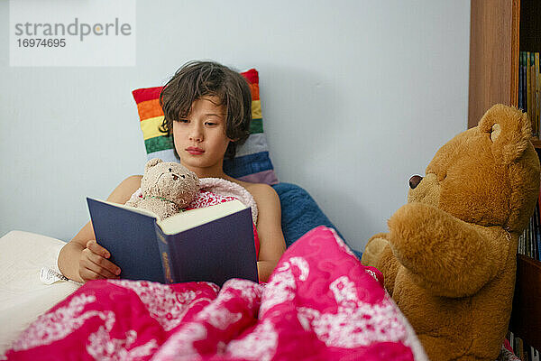Ein Junge liegt mit bunten Decken im Bett und liest mit Teddybären