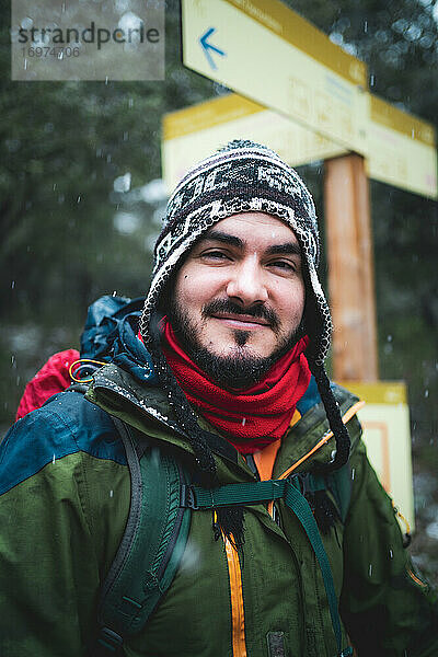 Mann lächelt mit Kälteschutz neben einem Trekking-Schild in die Kamera