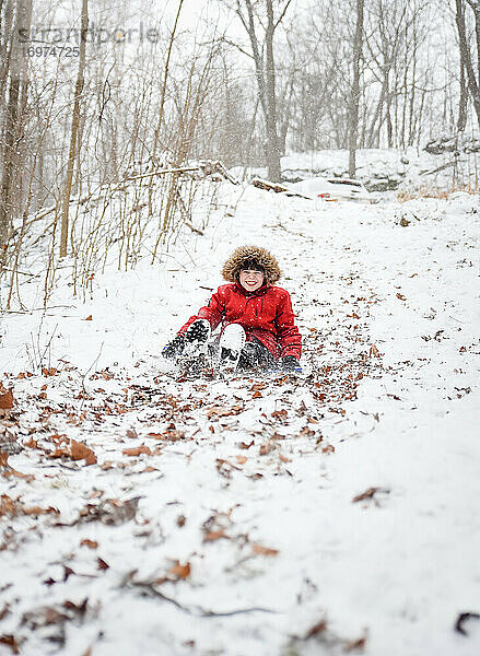 Fröhlicher Junge  der an einem verschneiten Wintertag einen Hügel im Wald hinunterschlittert.