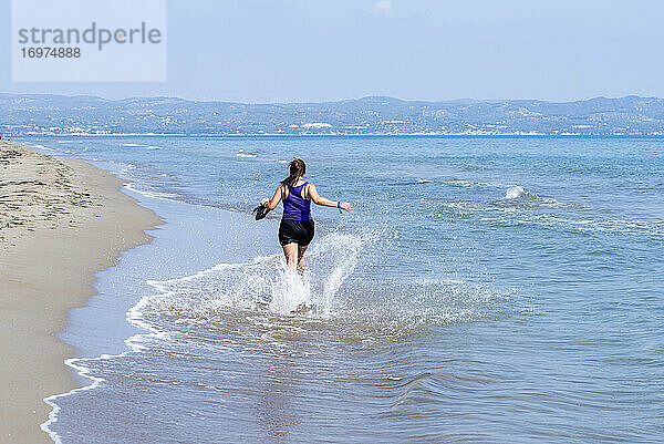 Rückenansicht einer Frau in Shorts  die am Meeresufer läuft und Wasser spritzt